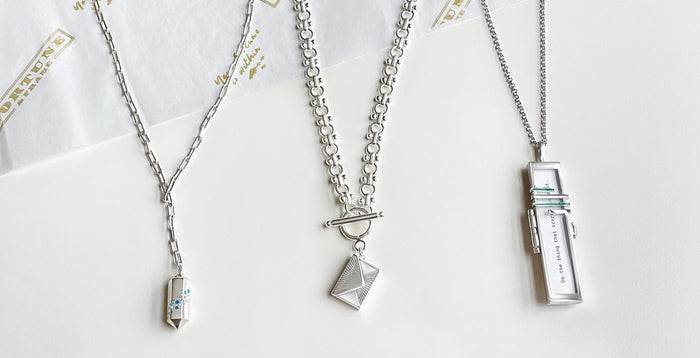 Silver Polygon Lariat Locket, Silver Love Letter Envelope Locket, and Silver Read Between The Lines Fortune Locket in a line, laying on top of Fortune & Frame tissue paper